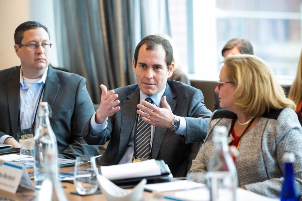 Three people in frame. Two men in the middle and left side, both wearing suits. Woman on the right wearing a tweed blazer.
