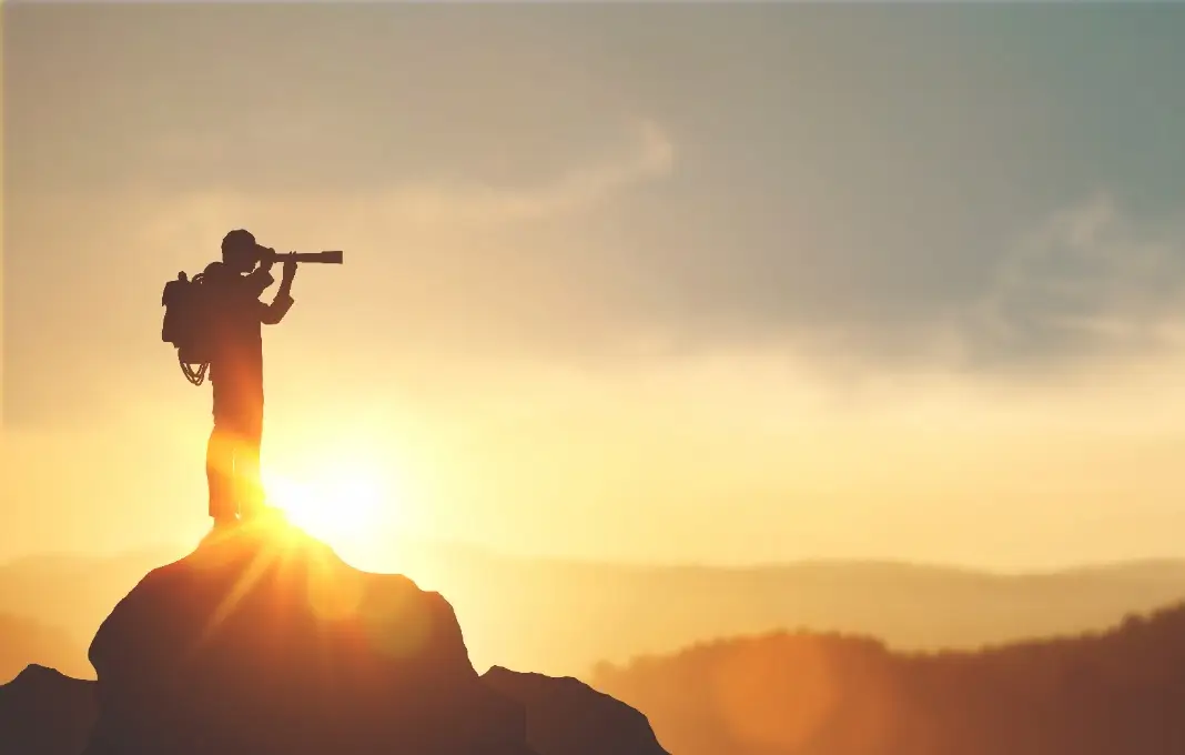 Silhouette of man holding binoculars on mountain peak against bright sunlight