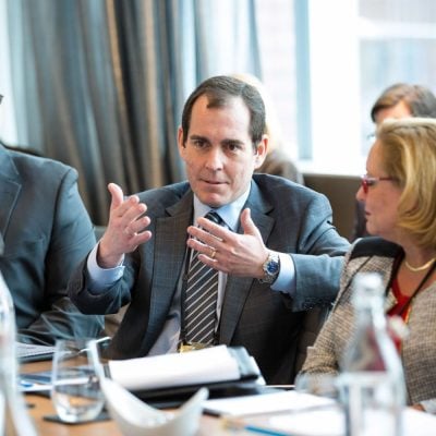 Three people in frame. Two men in the middle and left side, both wearing suits. Woman on the right wearing a tweed blazer.