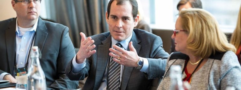 Three people in frame. Two men in the middle and left side, both wearing suits. Woman on the right wearing a tweed blazer.